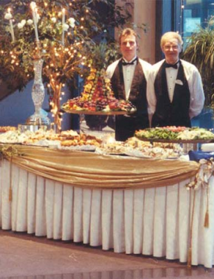 Two well-dressed servers in tuxedos stand proudly in front of a delectable buffet