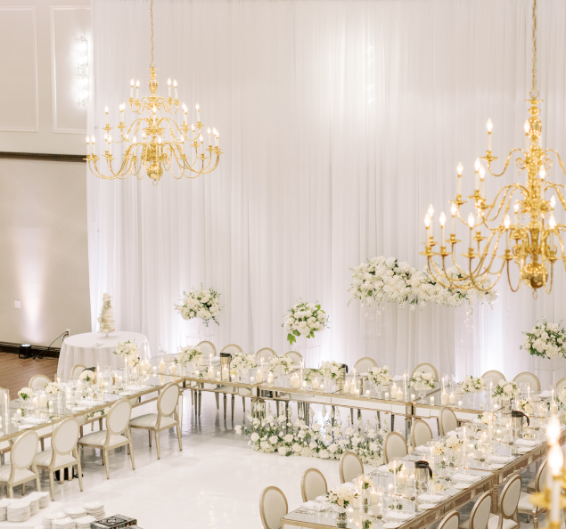 An elegant all-white ballroom adorned with exquisite gold chandeliers and tastefully arranged white and green flowers