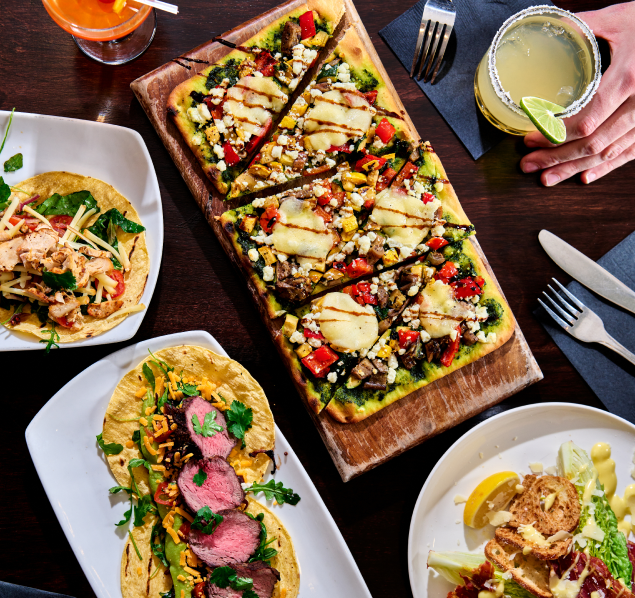 A mouthwatering display of a plate filled with delicious tacos, each topped with colorful and flavorful ingredients. Next to it, a scrumptious flatbread pizza tantalizes with its savory toppings, with a hand holding a refreshing drink, inviting you to sip and enjoy
